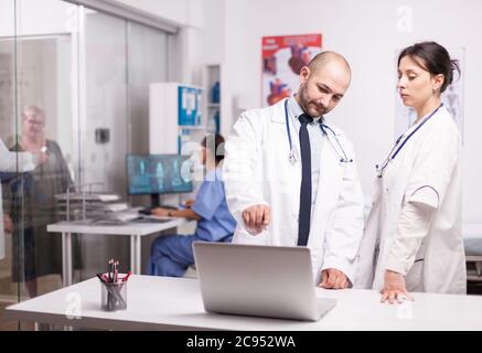 I giovani medici puntano allo schermo del portatile in ufficio dell'ospedale vestito con cappotto bianco. Anziana donna anziana e medica nel corridoio clinico che parla di prescrizione di malattia. Infermiere in uniforme blu che lavora sul computer. Foto Stock