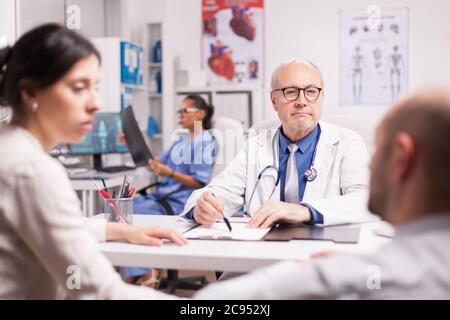 Medico senior durante la consultazione con un giovane in sedia a rotelle, accompagnato da moglie. Medico con rivestimento bianco e stetoscopio e infermiere che tiene l'immagine radiografica. Foto Stock