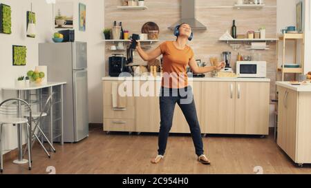 Giovane bella donna che balla mentre ascolta la musica con le cuffie blu  senza filo in cucina. Energica, positiva, felice, divertente e simpatica  casalinga che danzano da sola in casa. Intrattenimento e