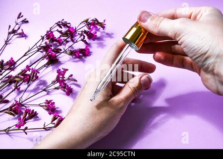 Donna gocciolando olio essenziale rosa a mano contro fiori sfocati su sfondo rosa, closeup, asciugamani bianchi spa, trattamento di bellezza, cura della pelle delle mani Foto Stock