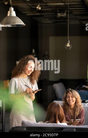 Giovane donna dai capelli ricci focalizzati immersa nella digitazione dei social media sul computer portatile mentre interagisce online in un bar Foto Stock