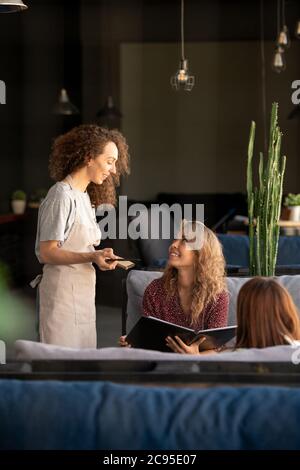 Giovane donna dai capelli ricci focalizzati immersa nella digitazione dei social media sul computer portatile mentre interagisce online in un bar Foto Stock