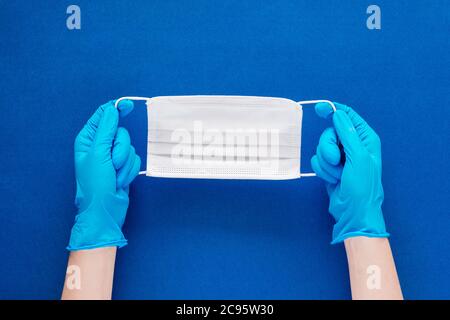 Foto in stock delle mani di una donna con guanti blu che tengono una maschera facciale su uno sfondo blu testurizzato Foto Stock