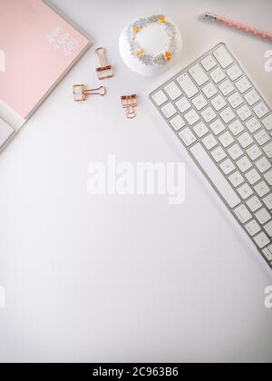 Scrivania da ufficio bianca con notebook rosa, tastiera bianca, forniture per ufficio e cupcake decorato. Disposizione piatta con spazio per la copia. Foto Stock