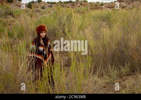 Giovane donna ispanica vestita come un trapezio in un poncho e cappello di pelliccia escursioni attraverso il paesaggio desertico con erba alta nella Ojito Wilderness, New Mexico Foto Stock