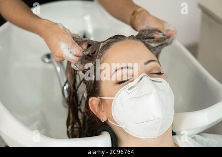 Giovane ragazza con lunghi capelli marroni, che si inzuppano lavando la testa nel parrucchiere. Distanza sociale. Uso della maschera facciale. Primo piano Foto Stock