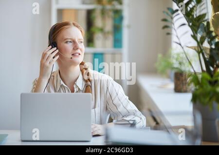 Donna dai capelli rossi in cuffia seduta sul suo posto di lavoro che lavora nel call center Foto Stock