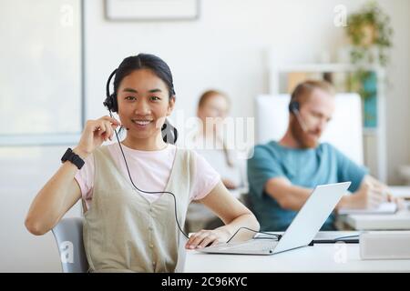 Ritratto della giovane donna asiatica in cuffia sorridente alla fotocamera mentre lavora sul suo posto di lavoro con un computer portatile Foto Stock