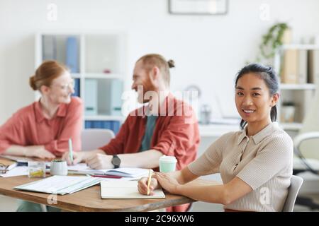 Ritratto di giovane donna asiatica sorridente alla macchina fotografica mentre si siede al tavolo con altri uomini d'affari al meeting in ufficio Foto Stock