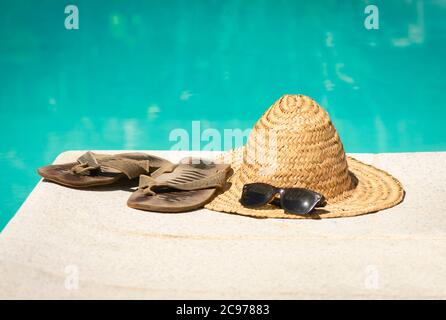 Cappello di paglia d'epoca con occhiali da sole e infradito al bordo di una piscina. Foto Stock