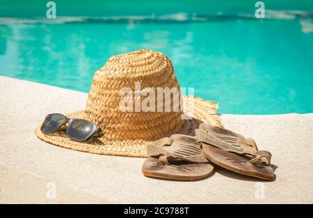 Cappello di paglia d'epoca con occhiali da sole e infradito al bordo di una piscina. Foto Stock