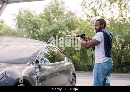 African elegante giovane uomo messo a fuoco in vestiti casual che lavano la sua moderna auto elettrica con una pistola ad acqua sulla stazione self-service di lavaggio auto all'aperto Foto Stock