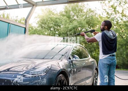 African elegante giovane uomo messo a fuoco in vestiti casual che lavano la sua moderna auto elettrica con una pistola ad acqua sulla stazione self-service di lavaggio auto all'aperto Foto Stock