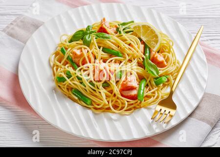 Deliziosa pasta al salmone con fagioli verdi e salsa al burro all'aglio con basilico fresco sulla parte superiore e limone servito su un piatto bianco con forchetta su un legno bianco Foto Stock