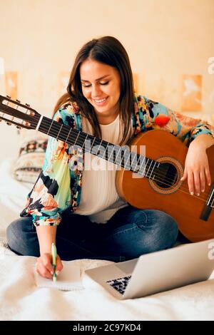 Crop musicista femminile con chitarra acustica e portatile che prende appunti mentre si siede a letto e scrivere canzone a casa Foto Stock