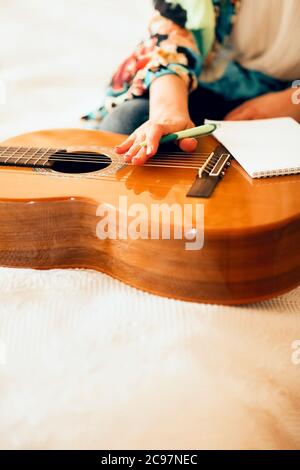 Crop musicista femminile con chitarra acustica e portatile che prende appunti mentre si siede a letto e scrivere canzone a casa Foto Stock