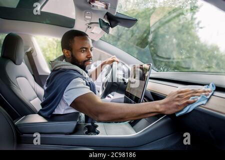 Giovane bell'uomo africano sorridente che pulisce la console dell'auto e il pannello di controllo con un panno in microfibra. Concetto di autolavaggio e dettaglio al autolavaggio Foto Stock