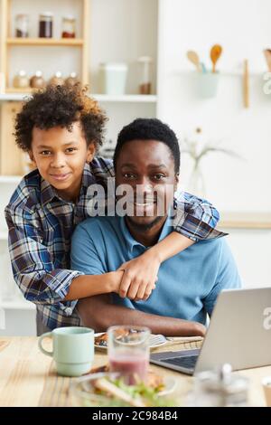 Ritratto di felice padre e figlio sorridenti alla macchina fotografica mentre si siede al tavolo con il computer portatile in cucina Foto Stock