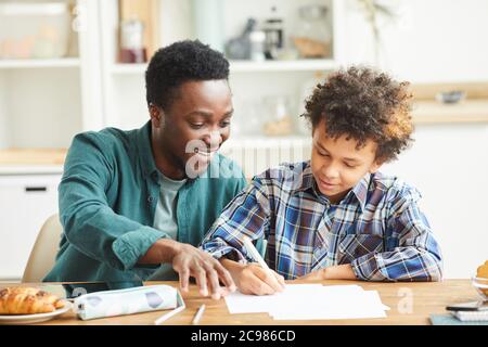 Africano sorridente padre che aiuta suo figlio a fare il suo lavoro che sedevano al tavolo nella stanza Foto Stock
