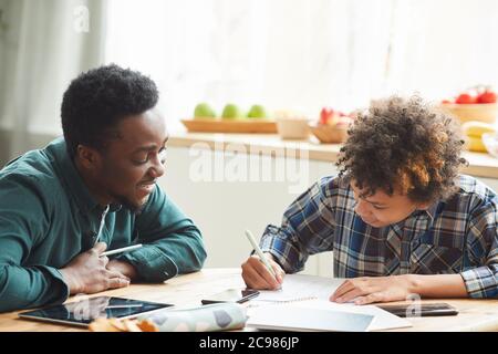 Padre africano aiutando suo figlio a studiare durante l'educazione online a casa l'uomo che spiega il materiale mentre il ragazzo fa appunti in notebook Foto Stock