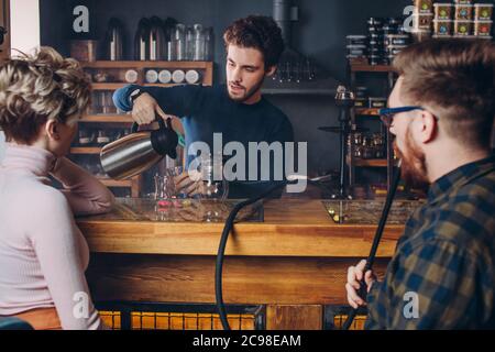 giovane ragazzo bearded attraente che serve una coppia. primo piano sparato. Barman caucasica preparazione di bevande per i visitatori Foto Stock