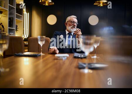 Uomo anziano dai capelli grigi che tiene e fuma e-sigaretta in un ristorante ricco. IQOS, tecnologie moderne, smetta di fumare Foto Stock