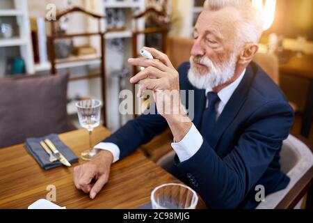 Elegante uomo anziano in suite fumo calore-non-bruciare la tecnologia del prodotto del tabacco sedersi nel ristorante, tenendo la sigaretta nella mano destra Foto Stock