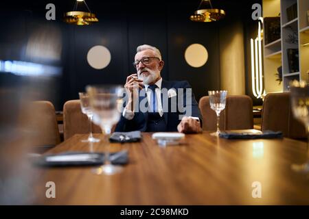 Uomo anziano dai capelli grigi che tiene e fuma e-sigaretta in un ristorante ricco. IQOS, tecnologie moderne, smetta di fumare Foto Stock