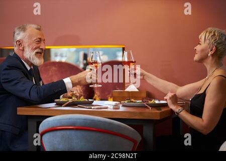 Coppia elegante senior sedersi al ristorante e clink bicchieri con champagne, festeggiare. Uomo dai capelli grigi in tuxedo e donna caucasica in nero dr Foto Stock