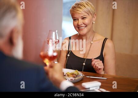 Donna caucasica anziana in abito nero, cibo raffinato per cena con l'uomo in un elegante ristorante. Femmina alzò un bicchiere e andando a bere. Vista posteriore sul marito Foto Stock