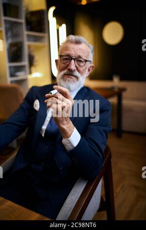 Bell'uomo anziano con capelli grigi in bicchieri in abito fumo IQOS in ristorante, esplorare la sigaretta tenendo in mano Foto Stock