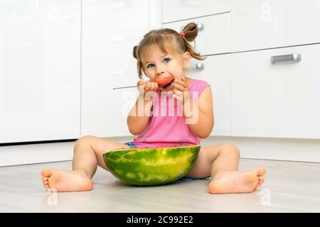 bambina di 4 anni con appetito mangia anguria con un cucchiaio seduto sul pavimento a casa ed è felice Foto Stock