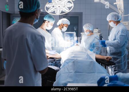 Serio chirurgo maschile abile in piedi nella sala operatoria e prendendo uno strumento, mentre gli altri membri della squadra lo assistono. Foto Stock