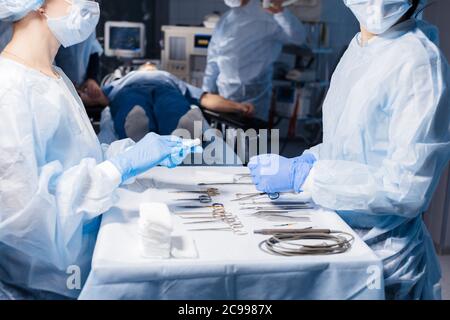 Due diverse infermiere che prendono strumento chirurgico per gruppo di chirurghi in background operating patient in sala operatoria,primo piano . Foto Stock