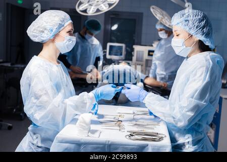 Due diverse infermieri femminili che assumono strumenti chirurgici per il gruppo di chirurghi in background operating patient in sala operatoria. Foto Stock