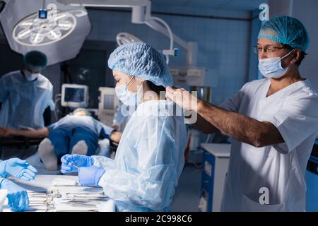 Chirurgia, medicina e concetto di persone - medico maschile che assiste il chirurgo femminile e la aiuta con i guanti e l'usura protettiva in sala operatoria all'Hospi Foto Stock