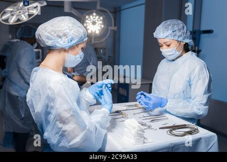 Due giovani asiatici e caucasici assistenti femminili preparare strumenti chirurgici e utensili disposti su una tavola per un intervento chirurgico, fornendo tutte le necessarie eq Foto Stock