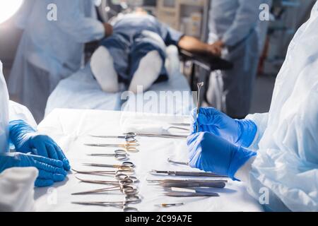 Concentrarsi su strumenti e strumenti chirurgici e sullo sfondo irriconoscibile paziente maschile giacente sul coach e squadra di chirurgia che lavorano insieme, con occupato Foto Stock