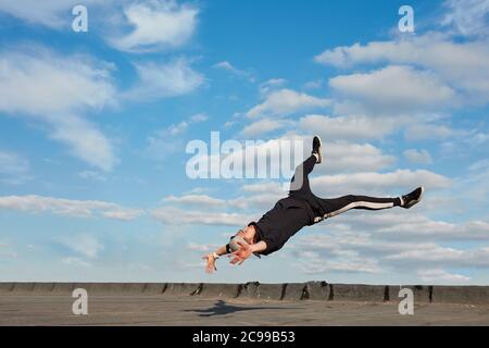 Ballerino asiatico hip-hop vestito con pantaloni sportivi neri, felpa saltando in aria, facendo un downrock acrobazie all'aperto sul tetto su sfondo blu cielo Foto Stock