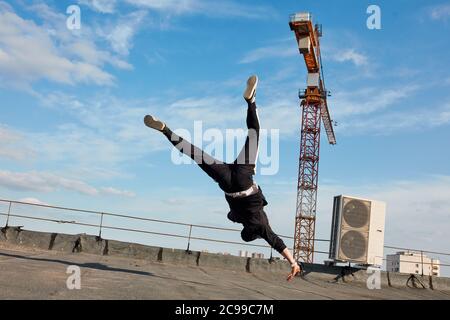 Ballerina di stile libero in vestiti neri che volano nell'aria facendo colpo che balla danzare la rottura danza sul tetto di costruzione con la gru di costruzione sullo sfondo- Foto Stock