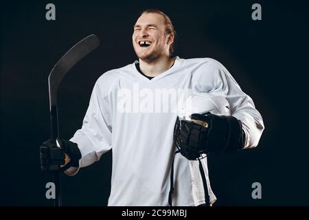 Felice bello giocatore di hockey con uno rotto dente anteriore ridere per fotocamera, in piedi con bastone in bianco uniforme, isolato su nero Foto Stock