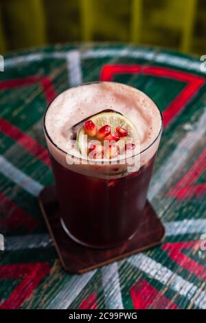 Un cocktail in un calice di highball con schiuma, cubetto di ghiaccio all'interno, guarnito con una ruota di lime e semi di melograno Foto Stock