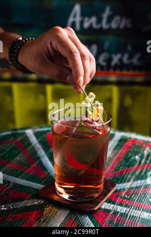 Un cocktail trasparente in un bicchiere di highball con un grande cubetto di ghiaccio. Una mano che mette gli orsi gommoso guarnire sopra una bevanda Foto Stock