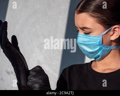 Femmina adolescente che si adatta su guanti in lattice nero in blu protettivo maschera medica viso. Occhi chiusi. Protezione antivirale durante la pandemia del coronavirus. Foto Stock