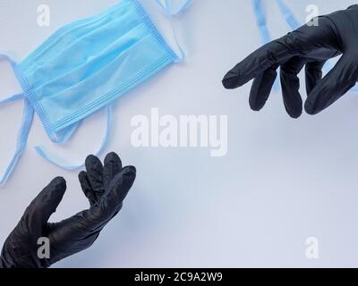 Due mani in guanti di lattice neri che si allungano l'una verso l'altra. Maschera medica protettiva blu su sfondo bianco. Protezione antivirale. Foto Stock