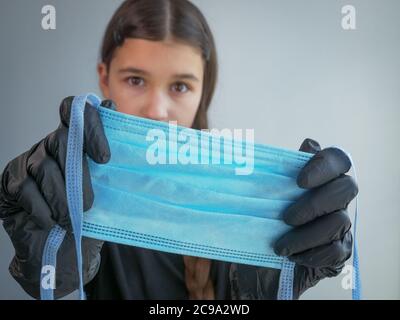 Femmina adolescente ragazza in nero guanti in lattice guardando nella macchina fotografica, tenendo e mostrando una maschera medica di protezione blu in primo piano. Antivirale Foto Stock