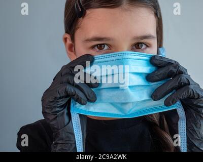 Femmina adolescente ragazza in nero guanti di lattice tenuta e montaggio su una maschera medica di protezione blu. Occhi chiusi. Protezione antivirale durante la covid. Foto Stock