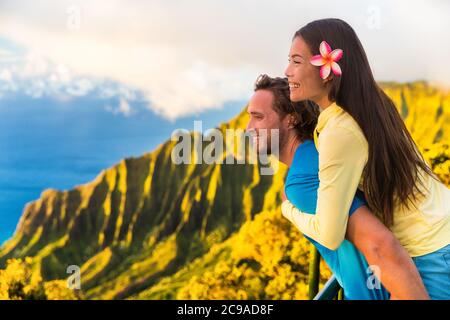 Viaggio coppia divertente piggyback a Na Pali Kauai Hawaii vacanza avventura. Felice giovane donna asiatica e uomo caucasico piggybacking a osservazione della natura in Foto Stock