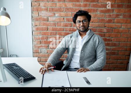 Positivo bell'uomo indiano seduto davanti al computer e in posa con la fotocamera. Uomo che riposa dopo il lavoro . primo piano foto Foto Stock