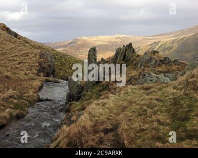 Cielo grigio sopra rocce scoscese e erba ruvida accanto a un torrente di montagna tumbling sopra Haweswater con lontane colline illuminate dal sole a Cumbria, Inghilterra, Regno Unito Foto Stock